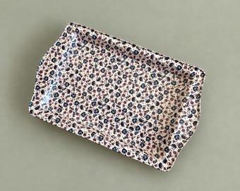 Puede incluir: Una bandeja de servicio rectangular con bordes ligeramente curvados, que presenta un patrón floral denso y repetitivo en tonos azul oscuro, granate y crema. La superficie tiene un acabado brillante y el diseño cubre toda la bandeja.