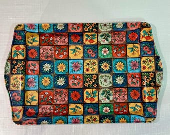 Puede incluir: Una bandeja de servicio rectangular con un patrón de cuadrícula vibrante de cuadrados individuales, cada uno decorado con distintas ilustraciones florales en tonos rojo, amarillo, azul, verde y naranja. La bandeja tiene bordes ligeramente elevados.