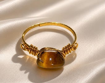 Puede incluir: Un anillo de alambre dorado con una piedra preciosa de ojo de tigre ovalada, lisa y pulida. El alambre de metal está enrollado alrededor de la banda a ambos lados de la piedra central, que muestra una característica chatoyancia marrón dorada.