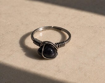 Puede incluir: Anillo plateado con una piedra azul oscuro en el centro. La piedra está sujeta por un diseño de alambre enrollado. El anillo se encuentra sobre un fondo neutro, con una sombra a un lado.