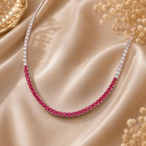 May include: A tennis necklace featuring a continuous row of round-cut stones. The design transitions from clear white stones on the upper sections to a vibrant segment of deep pink stones along the lower curve, set in a polished metallic frame.
