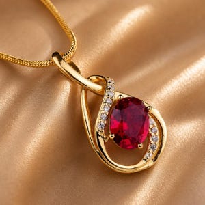 May include: A gold-colored pendant necklace featuring a large, oval-cut ruby gemstone. The ruby is surrounded by a halo of small, clear gemstones. The pendant hangs from a delicate gold chain, creating an elegant and eye-catching piece of jewelry.