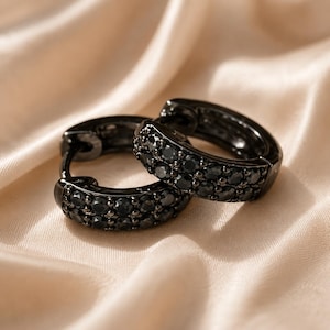 May include: A pair of small hoop earrings crafted from black metal, each featuring two parallel rows of embedded round black stones that catch the light. The earrings have a hinged closure mechanism and a polished finish on the metal surfaces.