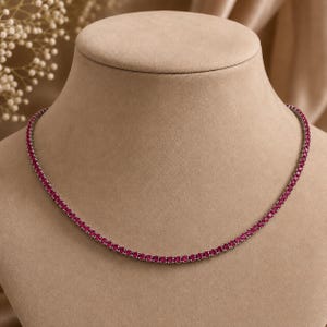 May include: A tennis necklace featuring a continuous row of small, round, vibrant pink-red gemstones set in a metallic frame. The necklace follows a circular path, showcasing uniform stones that create a consistent, sparkling line of color.