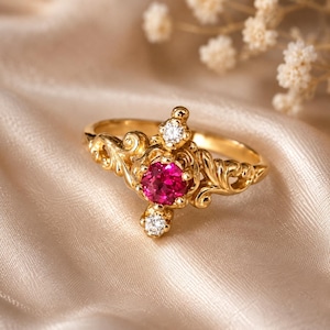 May include: A gold ring with a central, round, pink gemstone and two smaller white gemstones. The ring features ornate, swirling filigree designs. The ring is set against a soft, neutral background.