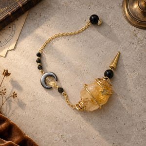 May include: A pendulum featuring a raw yellow citrine crystal wrapped in gold-tone wire with a small star charm. The chain includes black beads, a dark crescent moon charm, and a gold-tone metal spike tip at the end of the crystal point.