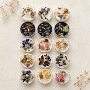May include: Fifteen round candles arranged in a grid, each topped with a unique combination of dried flowers, herbs, star anise, and various polished gemstones. The candles feature white or black wax bases and a single central cotton wick.