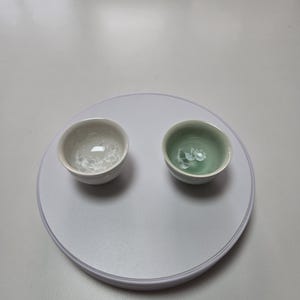 Może przedstawiać: Dwie małe, okrągłe ceramiczne filiżanki do herbaty. Jedna filiżanka jest w kolorze złamanej bieli z teksturowanym wnętrzem, a druga jest jasnozielona. Obie filiżanki znajdują się na białej, okrągłej powierzchni.