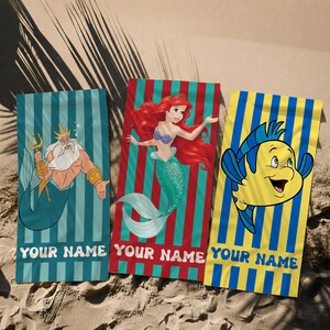 May include: Three beach towels with cartoon characters on a sandy surface. One towel features King Triton, another Ariel, and the third Flounder. Each towel has vertical stripes and the text "YOUR NAME" at the bottom.