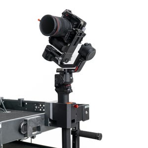 May include: A black DJI RS4 camera gimbal stabiliser mounted on a vertical support structure. The gimbal holds a professional camera with a large lens attached. The device features a black finish with red accents and the text RS4 clearly visible.