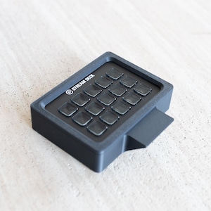 Pode incluir: Um Stream Deck preto com 15 botões quadrados transparentes. O dispositivo tem uma forma retangular com uma pequena extensão lateral angular. O texto "STREAM DECK" está impresso em branco na parte superior.