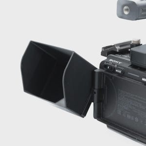 May include: A black camera sunshade attached to a black camera body. The sunshade is rectangular and angled, designed to block sunlight. The camera has a "SONY" logo and various buttons and dials.