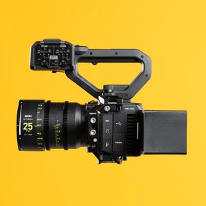 May include: A professional black video camera with a large lens and a handle. The lens has the text "25" and "Athena" printed on it. The camera has a flip-out screen and various buttons and ports. The background is yellow.