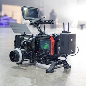 Puede incluir: Una cámara de cine profesional negra con asa superior, matte box de fibra de carbono, rueda de enfoque lateral, batería trasera con dos antenas y un monitor superior que muestra una interfaz digital con tonos de color verde.
