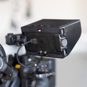 Pode incluir: Um dispositivo eletrônico preto com um botão giratório e botões cinza, conectado a uma câmera. O dispositivo tem uma luz vermelha acesa na parte superior e um cabo USB conectado. O dispositivo é montado em um sistema de câmera maior.