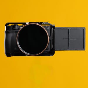 Puede incluir: Una cámara digital negra con un objetivo circular grande y un borde de color cobre. El cuerpo de la cámara está dentro de una jaula de metal negro y tiene una pantalla rectangular lateral con el texto en relieve Nikon RED en el panel.