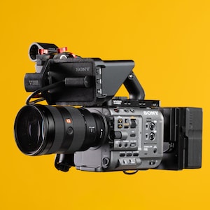 May include: A Sony FX6 cinema camera with a large lens and accessories, in matte black. The camera is set against a bright yellow background, highlighting its design and features. The camera is designed for professional use.
