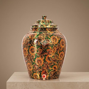 Puede incluir: Un dispensador de agua decorativo con tapa y un diseño vibrante y ornamentado. El dispensador de cerámica presenta un patrón floral verde y naranja, un grifo y una tapa a juego. El dispensador está colocado sobre una superficie clara.