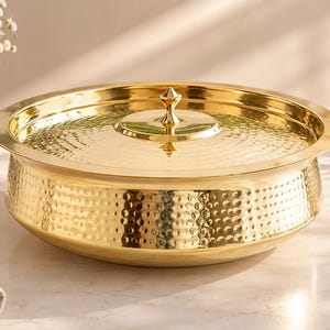May include: A round, gold-colored metal serving dish with a matching lid. The exterior of the bowl and the top of the lid feature a consistent hammered texture. The lid includes a small, pointed central handle for lifting the cover.