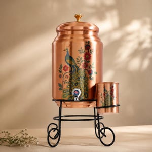 May include: A copper water dispenser with a lid and a spigot, featuring a colorful peacock and floral print. The dispenser sits on a black metal stand with a matching copper cup attached to the side, also decorated with a peacock and floral design.