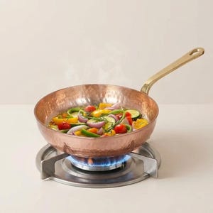 Pode incluir: Uma frigideira cor de cobre com uma alça de latão, cheia de vegetais coloridos, está em um fogão a gás com uma chama azul. A textura martelada da panela e os vegetais vibrantes criam uma cena de cozinha visualmente atraente.