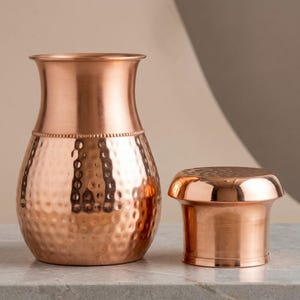 May include: A copper-colored vase and a small container. The vase has a wide base and a narrow neck, with a hammered texture on the body. The container has a flat top and a cylindrical body. Both items are made of shiny copper.