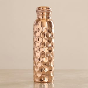May include: A cylindrical copper water bottle featuring a distinct hammered texture across its entire surface. The bottle has a smooth, rounded neck and a matching screw-top lid, showcasing a metallic rose-gold finish with reflective highlights.