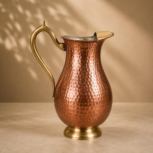 May include: A hammered copper pitcher featuring a polished brass handle and a matching brass base. The vessel has a wide, rounded body that tapers towards a flared spout, with a metallic finish that reflects light across its textured surface.