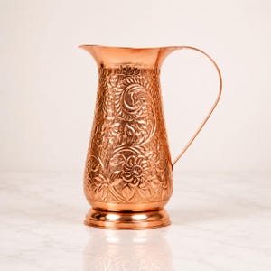 May include: A polished copper pitcher featuring an intricate embossed floral pattern covering the exterior surface. The vessel has a wide, flared rim, a tapered body, a sturdy circular base, and a single curved handle extending from the side.