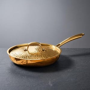 Op de afbeelding: Een goudkleurige koekenpan met bijpassende deksel. De pan heeft een lange handgreep en een gehamerde textuur op het kookoppervlak en deksel. De pan staat op een donkere ondergrond.