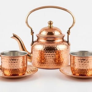 May include: A hammered copper tea set consisting of a kettle with a curved spout and a hinged handle, accompanied by two matching cups and saucers. Each piece features a textured, dimpled surface and a polished metallic finish.