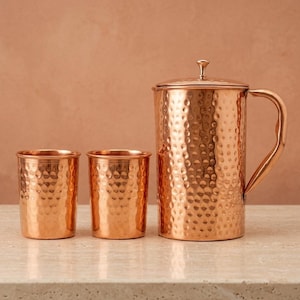 May include: A copper pitcher with a lid and handle, and two matching copper tumblers. The set has a hammered texture, reflecting light. The items are arranged on a light-coloured surface against a warm, neutral background.