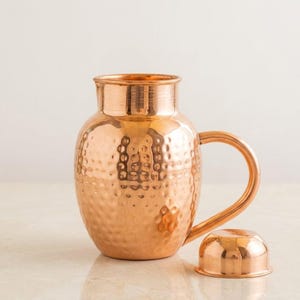 Puede incluir: Una jarra de cobre con textura martillada y asa curvada. La jarra tiene una tapa a juego y está diseñada para servir bebidas. El cobre tiene un tono marrón rojizo cálido y la superficie refleja la luz.