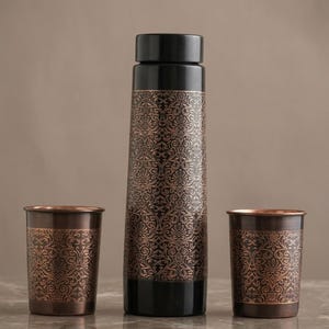 Puede incluir: Una botella de agua cilíndrica alta con tapa y base negras sólidas, con un intrincado patrón de damasco de color cobre en el cuerpo, acompañada de dos vasos a juego con el mismo diseño de damasco de cobre y bordes metálicos oscuros.