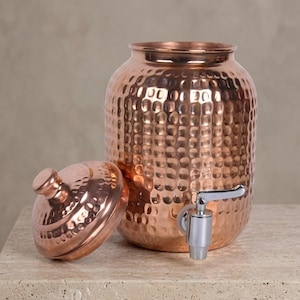 May include: A copper-coloured beverage dispenser with a hammered texture. The dispenser has a spigot and a lid. The lid is off to the side. The dispenser is on a light-coloured surface.