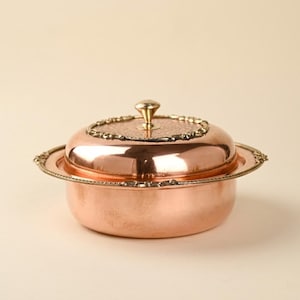 May include: A copper-colored butter dish with a lid and a decorative rim. The lid has a small, round handle and a detailed border. The dish sits on a matching base, creating a cohesive and elegant design. The overall aesthetic is vintage and ornate.