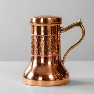May include: A copper-coloured pitcher with a lid and a gold-coloured handle. The body has a hammered texture, and the handle is curved. The pitcher has a wide base and a narrower top. The height is approximately 20 cm.