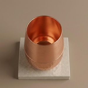 May include: A copper-colored stemless wine glass with a textured surface and a dotted band near the bottom. The glass sits on a square, light gray stone coaster. The background is a solid, neutral brown.
