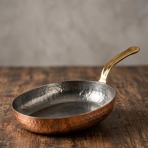 May include: A copper frying pan with a hammered finish and a brass handle. The pan has an oval shape and is set on a wooden surface. The handle curves upwards. The interior of the pan is a darker colour.