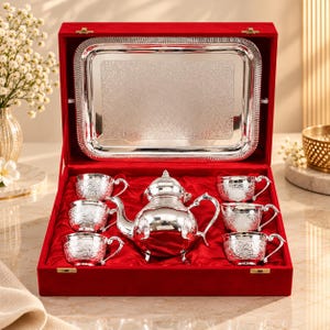 Op de afbeelding: Een zilveren theeservies met een theepot en vijf bijpassende kopjes met gedetailleerde bloemengravures, gepresenteerd in een met rood fluweel beklede doos. Een rechthoekig zilveren dienblad met een decoratieve rand zit in het deksel bevestigd.