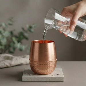 May include: A copper-colored stemless tumbler with a hammered texture on the lower half and a smooth upper half. Water is being poured into the glass from a clear pitcher. The glass sits on a square, light gray stone coaster.