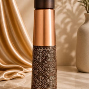 May include: A cylindrical water bottle featuring a smooth, polished copper upper section and a dark, matte-finished lower section. The lower portion is decorated with an intricate, repeating geometric floral pattern etched into the dark metal surface.
