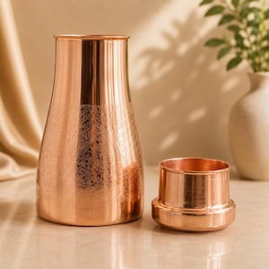 May include: A polished copper water pitcher with a tapered neck and a wide base, featuring an intricate floral etched pattern around the middle section. Beside the pitcher sits a matching short copper cup with a smooth, wide rim and base.