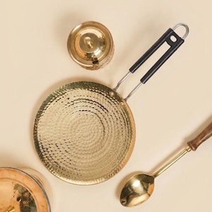 May include: A collection of brass kitchenware, including a hammered pan with a black handle, a small lidded container, and a spoon with a wooden handle. The items are arranged on a light beige surface.