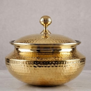 Op de afbeelding: Een goudkleurige, gehamerde metalen serveerschaal met bijpassend deksel. De kom heeft een afgeronde vorm met een licht uitlopende rand. Het deksel is bekroond met een decoratieve, bolvormige handgreep. Het oppervlak van de kom en het deksel heeft een getextureerd, kuiltjesachtig uiterlijk.