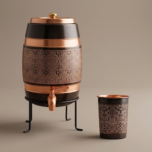 Puede incluir: Un dispensador de bebidas en forma de barril, negro y cobrizo, con un patrón floral decorativo. Tiene un grifo y se apoya en un soporte de metal negro. Hay una taza a juego al lado.