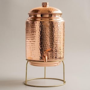 May include: A copper-colored beverage dispenser with a hammered texture, a spigot, and a lid. The dispenser sits on a gold-colored metal stand with a circular base. The dispenser is designed to hold and dispense beverages.