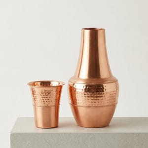 Puede incluir: Dos jarrones de metal color cobre. El jarrón más grande tiene un cuello alto y delgado y una base más ancha con una textura martillada. El jarrón más pequeño tiene forma de taza con una textura martillada. Ambos jarrones están sobre una superficie gris claro.