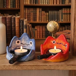 May include: Two ceramic candle holders shaped like stylized creatures with wide mouths, one in blue and one in red, each featuring a black handle and a lit tea light candle. The holders have simple painted eyes and wavy mouth lines.