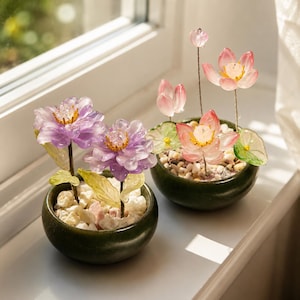 May include: Two small, dark green ceramic pots each hold a decorative flower arrangement. One features purple flowers, the other pink. Both have white pebbles and green leaves, creating a miniature garden scene.